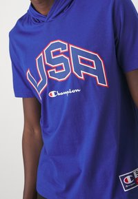 Niebieska bluza z krótkim rękawem z napisem "USA" w odważnych, czerwonych i białych literach. Logo "Champion" widoczne z przodu oraz metka po boku.