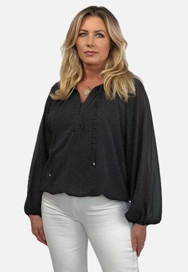 Czarna bluzka w białe kropki z dekoltem w serek, długimi rękawami balonowymi oraz detalem z troczkiem przy dekolcie. Dobrana z białymi dopasowanymi spodniami.