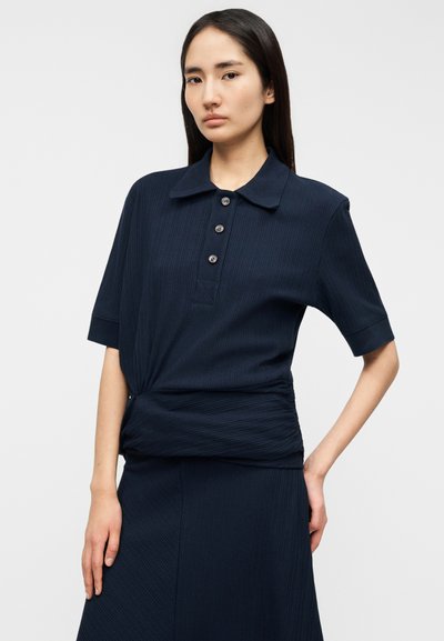 Μπλε ναυτικού κοντομάνικο πόλο με ραβδωτή υφή, κουμπιά και λεπτομέρεια με πιέτα στη μέση, συνδυασμένο με φούστα σε ίδιο χρώμα.