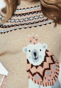 Pull en tricot beige avec un empiècement à motifs. Présente un graphique d'ours polaire vêtu d'un pull à motifs avec des touches de rose et de bleu.