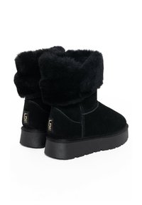 Botas de tobillo de ante negras con un suave collar plegable, superficie texturada, suela gruesa negra y un acento dorado del logo en el talón.