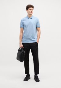 Gaiši zils polo krekls, melni bikses, melni sporta apavi un melna soma rokās. Gluda auduma, īsas piedurknes un apkakle.