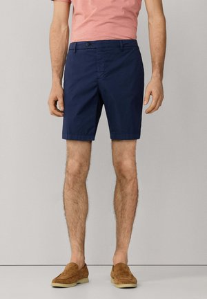 Man draagt marineblauwe korte broek, bruine suède loafers en een roze T-shirt, staand tegen een effen lichtgrijze achtergrond.