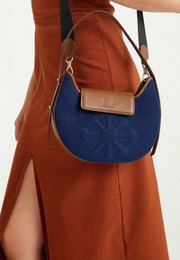 Dunkelblaue Canvas-Tasche mit braunen Lederelementen, geschwungene Form, Vorderklappe mit Verschluss und geprägtem floralem Muster. Verstellbarer Riemen.