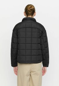 Jachetă puffer neagră, matlasată, cu un guler structurat, având cusături orizontale și o croială ușor scurtă, asociată cu pantaloni bej.