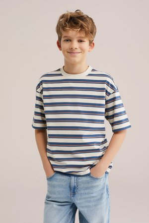 Smiling boy with tousled hair wearing a blue and white striped t-shirt and light blue jeans, hands in pockets, standing against plain background.