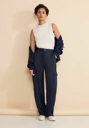 Pantaloni cargo blu con vestibilità rilassata, dotati di tasche laterali, abbinati a un top bianco senza maniche e a una giacca scura, su uno sfondo neutro.