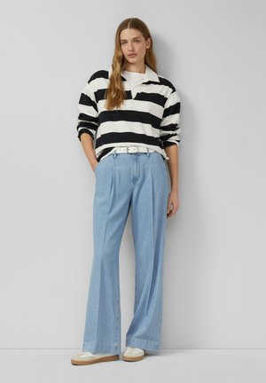 Woman wearing black and white striped long-sleeve shirt, light blue wide-leg jeans with white belt, and white sneakers, standing against gray wall.