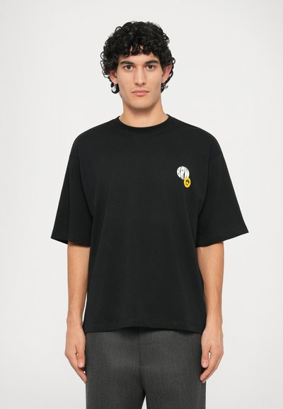 T-shirt nera in cotone con scollo rotondo, maniche corte e una grafica gialla con testo e un'emoticon sul petto.