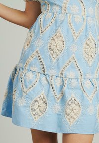 Helles blaues Baumwollkleid mit cremefarbenen Spitzenblumenmustern und ausgeschnittenen Details. Gestufter Schnitt mit gewelltem Saum und kurzen Ärmeln.