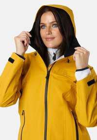 Gul vandtæt jakke med lynlås fortil, sorte detaljer og hætte. Har sidelommer og et lyst reflekterende logodesign.