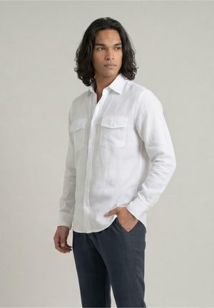 Man met schouderlang donker haar, gekleed in een wit overhemd met knopen en zakken en een donkere broek, staand met één hand in de zak.