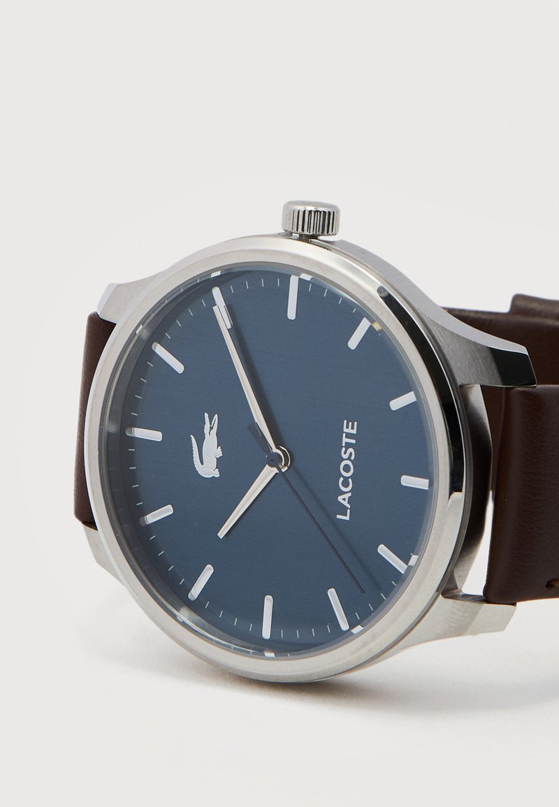 Runde silberne Uhr mit blauem Zifferblatt, weißen Stundenmarkierungen, Lacoste-Logo und braunem Lederarmband, zeigt die Zeit 7:55:45 an.