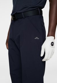 Marina golfbyxor med en stilren design, som har en logotyp på sidan. Ett svart texturerat bälte och en vit golfhandske med logotypdetaljer.