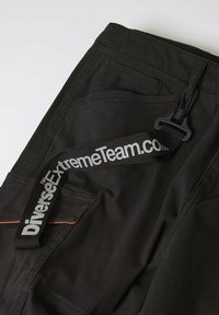 Czarne materiałowe spodnie z boczną kieszenią, z pomarańczowym wykończeniem i czarnym paskiem z napisem "DiverseExtremeTeam.com."