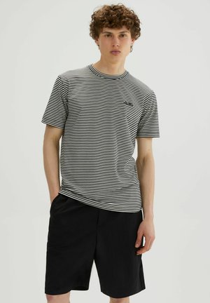 Joven con cabello rizado que lleva una camisa de manga corta a rayas blancas y negras y pantalones cortos negros, de pie contra un fondo claro y liso.