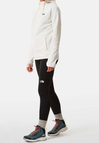 Felpa bianca in pile con zip frontale e due tasche laterali, abbinata a leggings neri con logo e scarponi da trekking blu con lacci rossi.