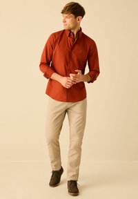 Cămașă button-up de culoare ruginiu, cu mâneci lungi, asortată cu chino bej și pantofi maro închis. Material neted și design ajustat.