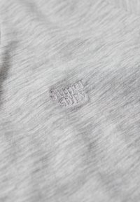 Lysegråt stof med subtil tekstur og lille hvidt broderet "Super Dry"-logo centreret i billedet.