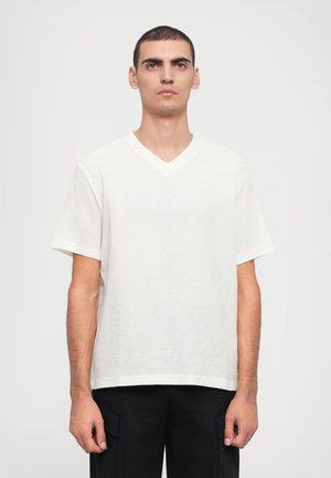 White short-sleeve V-neck shirt made of textured fabric with a subtle pattern, paired with black trousers, displayed against a plain background.