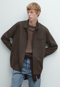 Bruine overhemd met kraag, een borstzak en een textuurstof, gelaagd over een bruine shirt en gecombineerd met blauwe spijkerbroek.