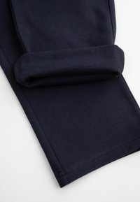 Granatowe spodnie z tkaniny o gładkiej fakturze, z podwiniętym mankietem. Po szwie wzdłuż krawędzi widać staranne przeszycia, podkreślające czyste linie.