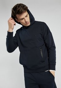 Granatowy hoodie z zamkiem błyskawicznym w przedniej kieszeni, miękka tkanina i ściągacze przy mankietach. Ma logo na klatce piersiowej oraz luźny krój.