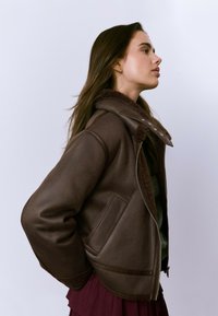 Veste en shearling marron avec fermeture éclair, grand col et poches latérales. Texture douce, coupe décontractée et détails de doublure en peluche très visibles.