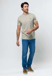 Čovjek stoji, nosi bež majicu kratkih rukava, plave traperice i smeđe tenisice, gledajući blago lijevo na jednobojnoj pozadini.