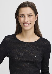Top lungo nero a maniche lunghe realizzato in pizzo con una texture a motivo rosa, dotato di un collo rotondo e di un design aderente.