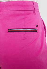 Pantalones de algodón rosa con un bolsillo de ojal lateral con detalles a rayas y una pequeña etiqueta de logo. Textura suave y diseño de corte regular.