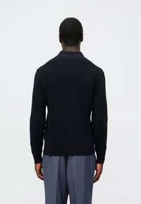 Tmavomodrý pletený polo sveter s dlhým rukávom a voľným strihom, s golierom a bočnými rozparkami, v kombinácii so svetlosivými nohavicami.