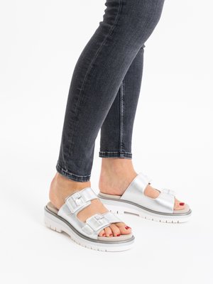 Füße in metallisch silbernen Sandalen mit Schnallen, dicken weißen Sohlen und dunklen engen Jeans, die auf einer weißen Oberfläche stehen.