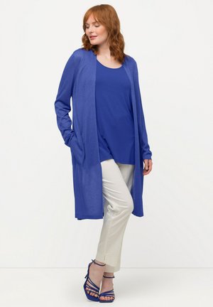 Langer, offen frontierter Cardigan in leuchtendem Blau mit langen Ärmeln, kombiniert mit einem passenden Top und cremefarbenen Hosen. Er zeichnet sich durch eine lockere Passform und zwei Taschen aus.