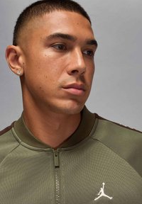 Olivgrön jacka med dragkedja framtill, ribbad krage och en vit Jumpman-logotyp. Jackan har en strukturerad yta och kontrasterande bruna detaljer.