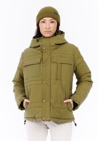 Persoon in een olijfgroene winterjas met capuchon en bijpassende beanie, staand met handen in de jaszakken tegen een witte achtergrond.