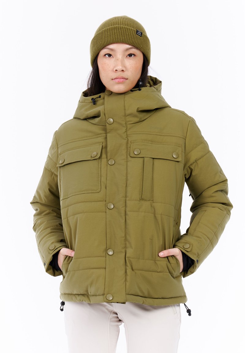 Persoon in een olijfgroene winterjas met capuchon en bijpassende beanie, staand met handen in de jaszakken tegen een witte achtergrond.