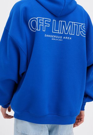 Sudadera con capucha azul con texto grande en blanco "OFF LIMITS" y texto más pequeño "DANGEROUS AREA BERLIN 2001" en la parte trasera, combinada con vaqueros de mezclilla claros.