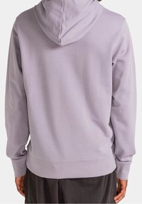 Persona che indossa un semplice hoodie viola chiaro visto da dietro, abbinato a pantaloni scuri su uno sfondo neutro.
