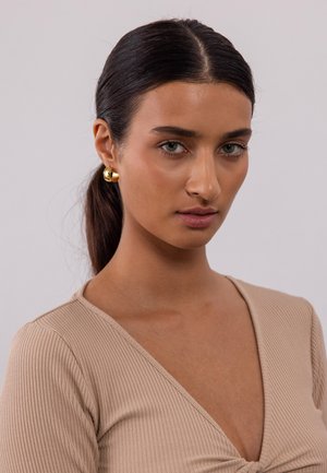 Beige, ribbelndes Langarmoberteil mit V-Ausschnitt und einem gedrehten Vorderdesign. Sichtbare goldene Stud-Ohrringe. Glattes Haar, neutraler Hintergrund.