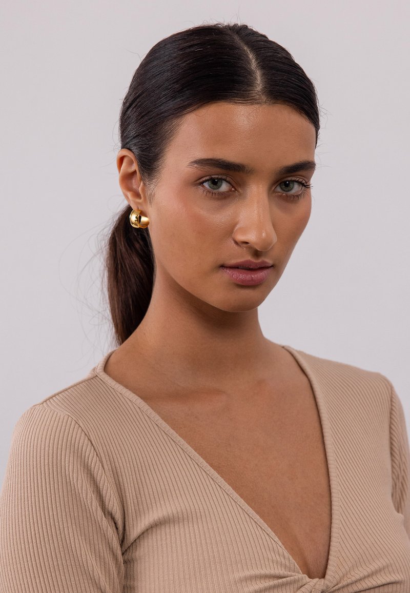 Top beige à manches longues en maille côtelée avec un décolleté en V, présentant un design avant torsadé. Boucles d'oreilles en or visibles. Cheveux lisses, fond neutre.