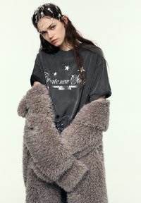 Grå oversized T-shirt med hvid grafisk tekst og stjerner, lagdelt under en fluffy grå frakke. Længere hår delvist dækket af et dekorativt hovedpynt.