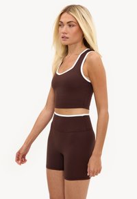 Femme blonde portant un crop top de sport marron et un short taille haute assorti avec une bordure blanche, debout devant un fond uni.