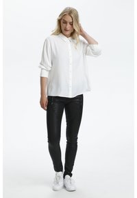 Chemise blanche à boutons avec manches retroussées, assortie à un pantalon noir slim avec des accents zippés. Portée avec des baskets blanches.