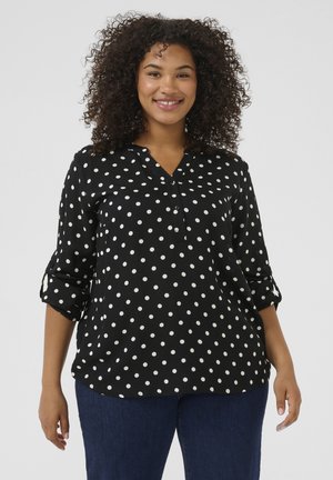 Blouse noire à pois avec un col à boutons, manches longues retroussées jusqu'aux coudes et coupe décontractée. Fabriquée dans un tissu lisse.