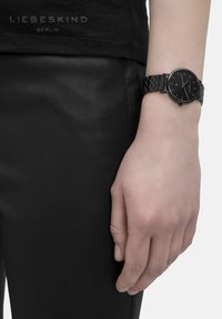 Schwarze runde Uhr mit einem glatten Gehäuse und einem strukturierten Metallarmband, getragen an einer Hand gegen schwarze Lederhosen und ein schwarzes Oberteil.