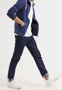 Sudadera navy con cremallera y cordones, camisa polo blanca, jeans azul oscuro y zapatillas blancas. Conjunto casual que presenta bolsillos laterales y puños doblados.