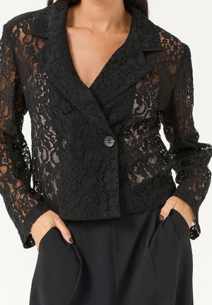 Schwarzer Spitzenblazer mit Blumenmustern, vorne mit Doppelknopf, getragen zu schwarzen Hosen, Hände in den Taschen, dunkler Nagellack sichtbar.