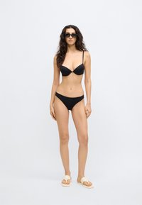 Σετ μαύρου μπικίνι με λεπτές ρυθμιζόμενες τιράντες και λεία υφή· το μοντέλο φορά μαύρα γυαλιά ηλίου και ανοιχτό μπεζ σανδάλια.