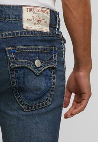 Vaqueros de mezclilla azul oscuro con un diseño distintivo en el bolsillo trasero de True Religion, con costuras azules en contraste y una etiqueta de marca prominente.